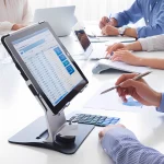 E-Rechnungssoftware Tablet auf einem Schreibtisch zeigt eine E-Rechnungssoftware mit Zahlenübersicht, während Personen im Hintergrund an Laptops arbeiten und Notizen machen. Symbolisiert die Bedeutung geeigneter Softwarelösungen für die E-Rechnungspflicht 2025.